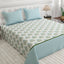 A neatly made bed in a bright bedroom setting, featuring a white bedspread with a repeating light blue and green floral block print, two matching light blue patterned pillows, and a light blue bed skirt with a white polka dot pattern.