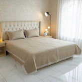 Beige bedspread on a bed in a well-lit bedroom with a lamp and curtains.