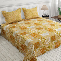 A patchwork quilted bedspread set in shades of yellow, orange, and beige with matching floral pillowcases, styled on a bed in a bright bedroom setting.