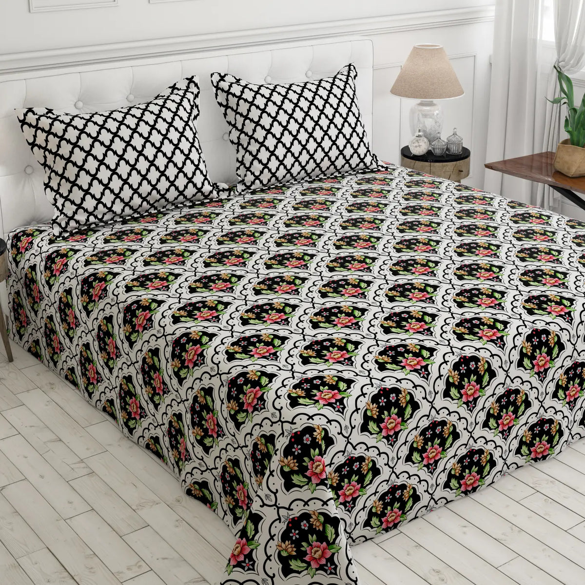 A bed dressed in a black and white patterned sheet set with matching pillowcases, featuring a repeating floral arch motif in pink, green, and yellow. The bed is styled in a bright bedroom with white paneled walls, a bedside lamp, and light wood flooring. Main image