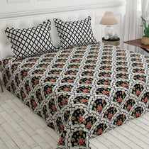 A bed dressed in a black and white patterned sheet set with matching pillowcases, featuring a repeating floral arch motif in pink, green, and yellow. The bed is styled in a bright bedroom with white paneled walls, a bedside lamp, and light wood flooring.