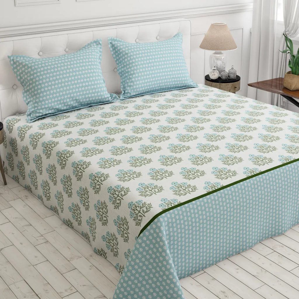 A neatly made bed in a bright bedroom setting, featuring a white bedspread with a repeating light blue and green floral block print, two matching light blue patterned pillows, and a light blue bed skirt with a white polka dot pattern.