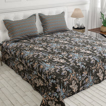 A bed dressed in a dark charcoal floral bedding set with matching patterned pillows, styled in a bright bedroom with a white tufted headboard, bedside lamp, and light wood floor.