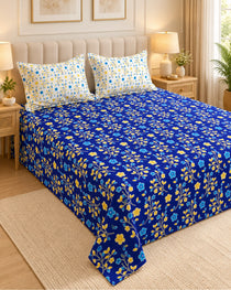 A blue floral bedspread set with two matching pillowcases on a bed in a styled bedroom. The bedspread features a vibrant royal blue base with a repeating pattern of yellow and light blue flowers and green stems.