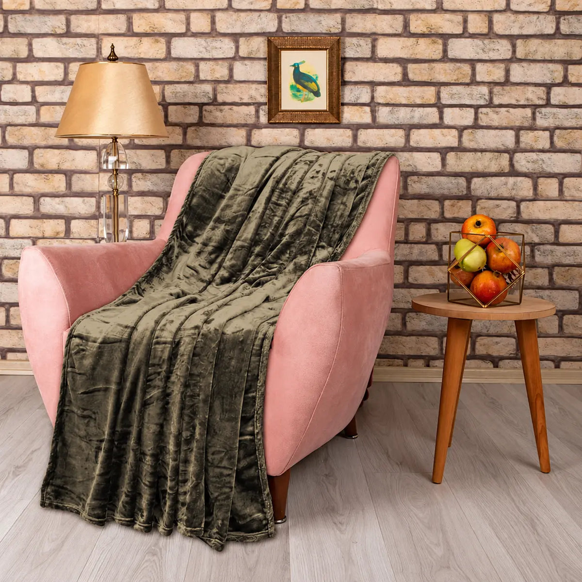 A plush olive green throw blanket is draped over a pink armchair in a styled living room setting with a brick wall, lamp, and side table. Main image