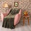 A plush olive green throw blanket is draped over a pink armchair in a styled living room setting with a brick wall, lamp, and side table.
