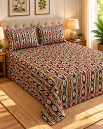 A bed dressed in a patterned beige comforter and matching pillow shams, styled in a warm bedroom with wooden nightstands, lamps, framed botanical wall art, and a potted plant.