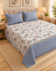 A neatly made bed in a warm bedroom setting, featuring a cream-colored floral print bedspread with blue and beige flowers, two matching blue striped pillows, and a blue striped bed skirt. The bed is styled with a beige tufted headboard, wooden nightstands with lit lamps, and a woven rug.