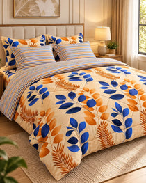 A bed dressed in a patterned comforter set with blue and orange leaf motifs on a cream background, matching striped pillows, and a striped folded sheet, shown in a warmly lit bedroom.