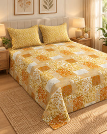 A bed dressed in a patchwork quilted bedspread and two matching pillowcases, featuring a yellow, orange, and white floral and geometric pattern, set in a warmly lit bedroom.