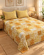 A bed dressed in a patchwork quilted bedspread and two matching pillowcases, featuring a yellow, orange, and white floral and geometric pattern, set in a warmly lit bedroom.